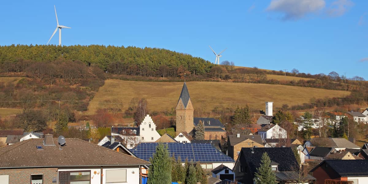 Blick über ein Eifeldorf mit Kirchturm, Häusern mit Solardächern und einem Hang mit Wald und Windrädern